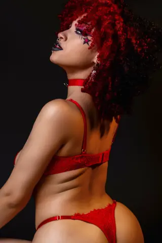 Woman with curly red hair in red lace lingerie posing against a dark background with a moody aesthetic.