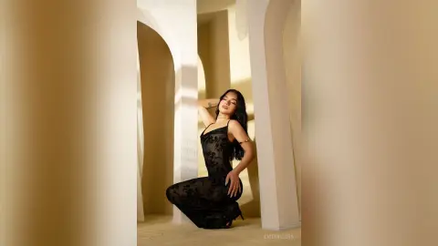 Elegant woman in a black lace dress posing in a sunlit minimalist white arched hallway.