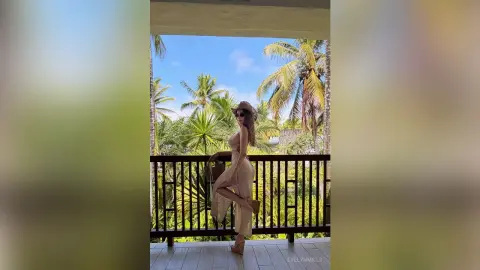 Stylish woman in a beige summer dress and sun hat posing on a tropical balcony overlooking palm trees.