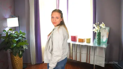 RubeWilliamson poses in a grey zip-up hoodie and blue jeans, looking back over her shoulder in a room with purple curtains and a silver dresser.