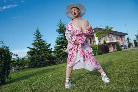 Elegant woman in a white and pink floral maxi dress and sun hat posing on a sunny green lawn.