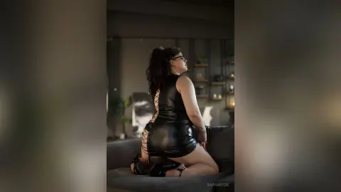 Curvy woman in a black leather mini dress sitting on a sofa in a moody, dimly lit modern living room.