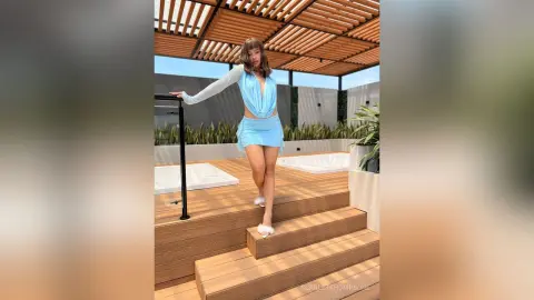 Young woman in a light blue summer outfit posing on a wooden deck under a pergola in bright sunlight.