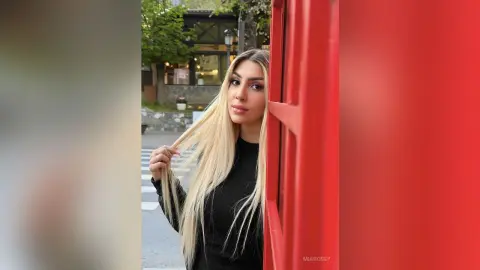 Blonde woman in a black long-sleeve top leaning against a bright red wall in an urban outdoor setting.