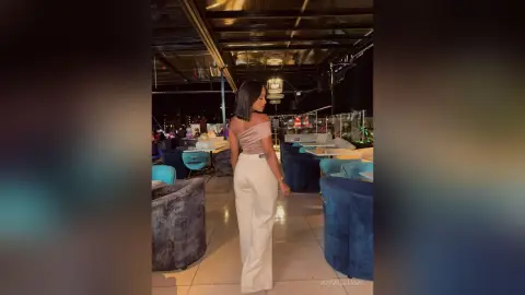 Elegant woman in a pink off-the-shoulder top and white trousers standing in a modern, dimly lit lounge.