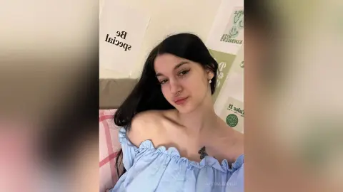 Young woman with long dark hair in a light blue off-the-shoulder top posing in a cozy indoor setting.