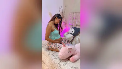 Pregnant woman in a light blue tank top relaxing on a bed with plush stuffed animals in a cozy bedroom.