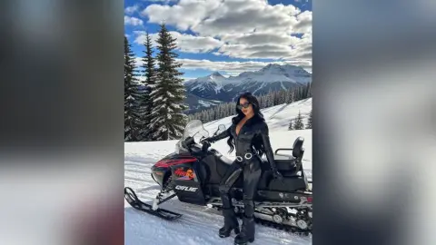 Stylish woman in black leather outfit posing with a snowmobile in a sunny, snowy mountain landscape.