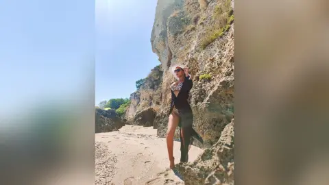 Stylish woman in a black outfit and sunglasses posing on a sunny rocky beach by a steep cliff.