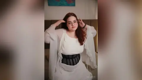 Young woman in white outfit and glasses posing thoughtfully in a cozy bedroom setting.