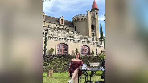 MiaBolson — Elegant woman in a burgundy dress sitting by a stone castle courtyard on a sunny day.