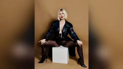 Blonde woman in black leather coat and leopard print tights posing confidently on a white pedestal.