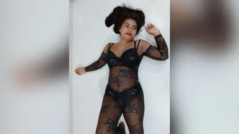Woman in a black lace bodysuit posing against a white wall in a moody, elegant style.