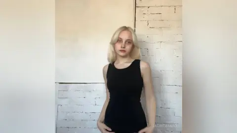 Blonde woman in a sleek black tank top standing against a white brick wall in a minimalist setting.