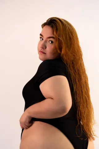 Curvy woman with long red hair wearing a black bodysuit against a white studio background.