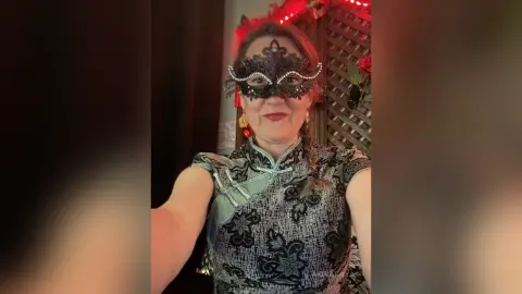 Woman in a black lace masquerade mask and patterned cheongsam dress posing in a moody, dimly lit room.