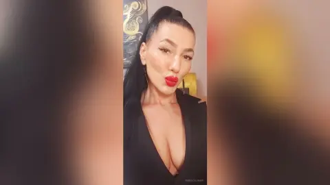 Glamorous woman with red lipstick and high ponytail wearing a black plunging dress in a warm indoor setting.