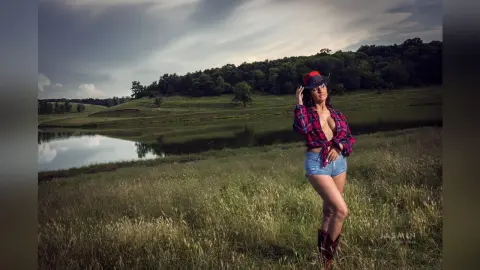 MissyJolie stands in a grassy field by a lake wearing a red plaid shirt and denim shorts.