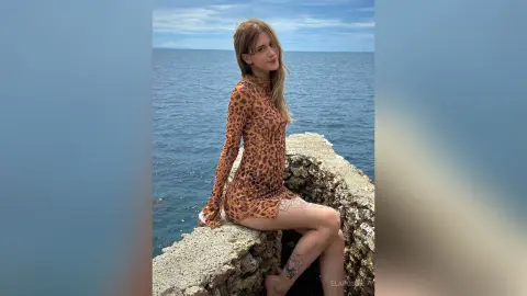Young woman in a leopard print long-sleeve mini dress sitting on rocks by the blue ocean under a clear sky.