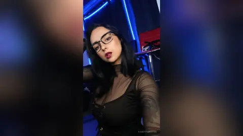 Young woman with glasses in a black sheer mesh top posing in a moody blue neon lit room.