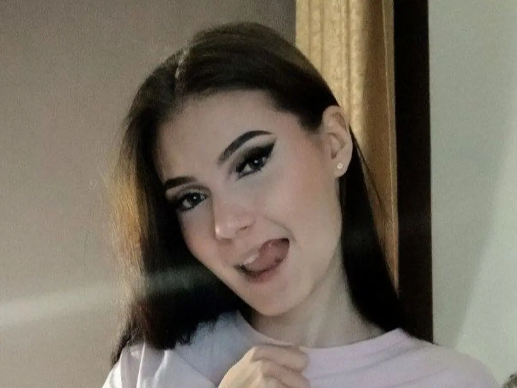 Young woman with long dark hair and winged eyeliner making a playful expression in a cozy indoor setting.