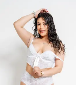 Smiling woman with wavy dark hair wearing white lace lingerie in a bright studio setting.