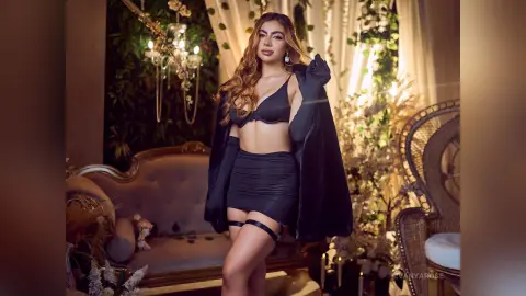 Glamorous woman in a black mini skirt and bra set posing in a lush, romantic indoor garden setting.