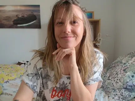 Smiling blonde woman in a white floral t-shirt relaxing on a bed in a cozy bedroom setting.