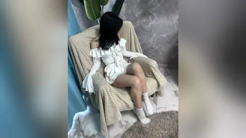 RittaMyers sits on a tan textured chair wearing a white off-the-shoulder ruffled outfit and white socks, looking away from the camera.