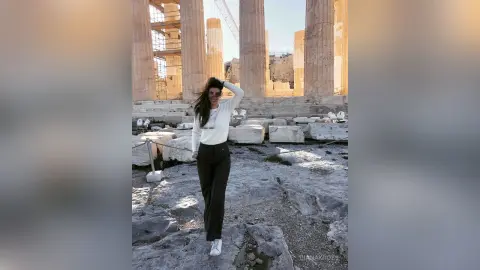 Smiling woman in white sweater and black trousers standing amidst ancient Greek ruins at the Acropolis.