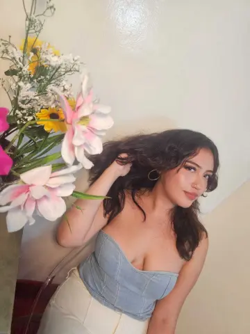 Young woman in a blue denim corset and white pants posing near pink flowers in a bright indoor setting.