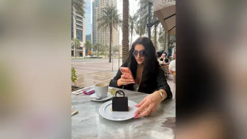 Stylish woman in a black blazer using her phone at an outdoor urban cafe with city skyscrapers in the background.