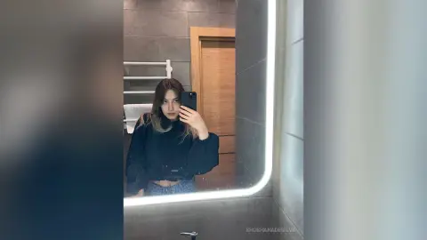 Young woman in a black sweatshirt taking a moody mirror selfie in a modern bathroom.