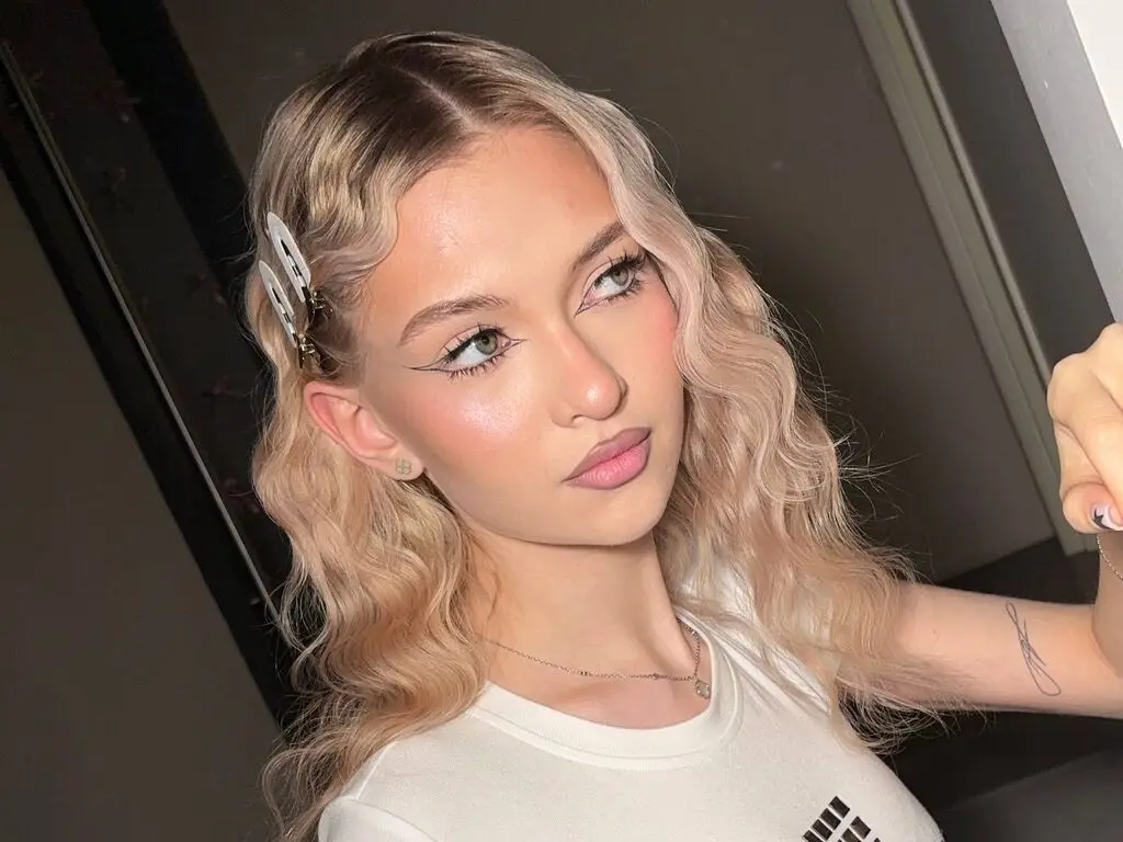 Young woman with wavy blonde hair and winged eyeliner wearing a white t-shirt in a soft indoor setting.