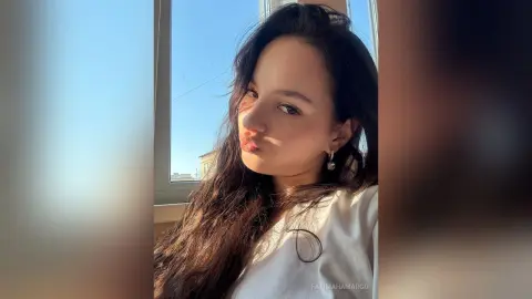 Young woman with long dark hair in a white top posing by a sunny window in soft natural light.