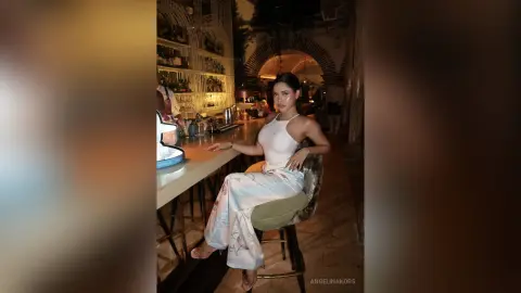 Elegant woman in a white halter top and floral trousers sitting at a dimly lit luxury bar.