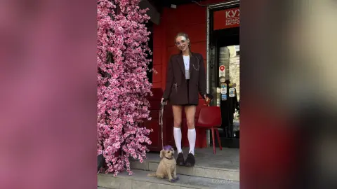 Stylish young woman in a brown blazer and white socks standing by pink flowers with her small dog.