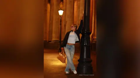 Stylish woman in jeans and white top leaning against a lamppost on a warm, moody city street at night.