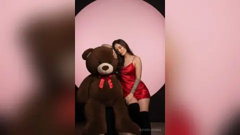 Young woman in a red satin slip dress leaning against a giant teddy bear in a dreamy pink studio setting.