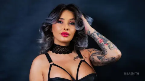 Tattooed woman with wavy hair wearing a black lace choker and lingerie against a dark moody background.