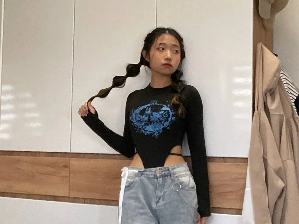 Young woman in a black graphic long-sleeve crop top and light denim jeans posing in a modern minimalist room.