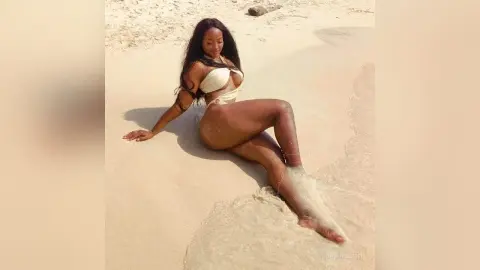 NohomiCambel reclines on a sandy beach, wearing a white bikini while exposing her bare skin and legs.