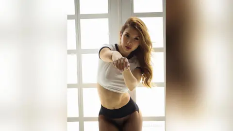 Athletic woman in a white crop top and black briefs posing confidently in a bright, sunlit room.