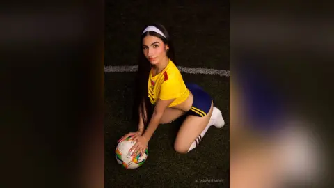 AlmaWhitmore poses on a grass field wearing a yellow crop top and blue shorts, holding a soccer ball while kneeling.
