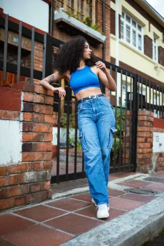 Curvy woman in blue crop top and baggy jeans leaning against a brick wall in a sunny urban setting.