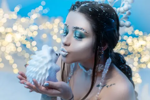 Winter queen with icy makeup and white outfit kissing a small snow hedgehog in a magical bokeh setting.