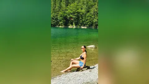 Woman in black bikini top and denim shorts sitting on a rock by a serene turquoise mountain lake.