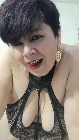 Smiling woman with short dark hair wearing a black mesh cutout bodysuit in a bright indoor setting.