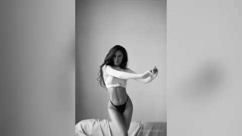 MilanaVolkova poses in a white long-sleeve crop top and black thong, showing her abs and hips against a plain grey background.
