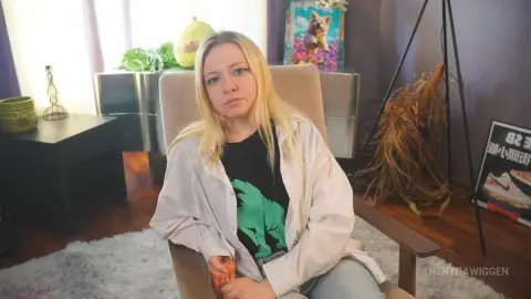Blonde woman in a black graphic tee and white shirt sitting in a cozy living room with a relaxed mood.